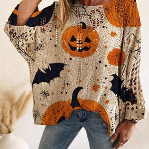 May include: A beige open-knit sweater featuring a Halloween-themed pattern with orange jack-o'-lanterns, black bats, and spiderwebs. The loose-fitting garment has long sleeves and a wide neckline, with the text NOXELLERATELIER printed at the bottom.