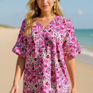 May include: A short-sleeved tunic top featuring a vibrant pink, magenta, and white abstract patterned print with a V-neckline and a slightly longer hemline in the back. The garment has a relaxed fit with wide sleeves and a subtle side slit detail.