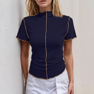 May include: A dark navy blue short-sleeve top featuring a mock neck and contrasting tan exposed stitching along the center front, shoulders, and sleeve hems. The garment has a lettuce-edge finish on the collar, sleeves, and bottom hem.