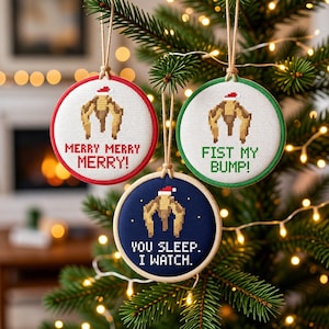 Può includere: Tre decorazioni circolari a punto croce appese con spago. Quella con bordo rosso dice MERRY MERRY MERRY!, quella verde FIST MY BUMP! e quella blu YOU SLEEP. I WATCH. Ognuna raffigura una creatura pixelata che indossa un cappello da Babbo Natale.