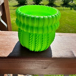 May include: A bright neon green plant pot featuring a textured exterior with a raised vertical cable knit pattern. The cylindrical container has a wide, reinforced rim and a deep interior, showcasing a vibrant, solid colour throughout the entire piece.