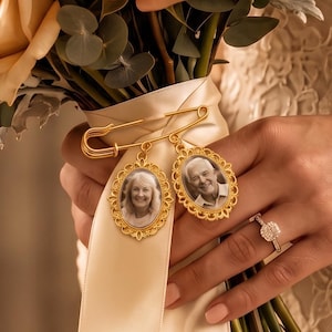 Pode incluir: Um broche de alfinete de segurança em tom dourado com dois pingentes de foto ovais com bordas recortadas ornamentadas. Os pingentes exibem retratos em preto e branco de uma mulher idosa e um homem idoso, presos a uma fita de cetim creme.