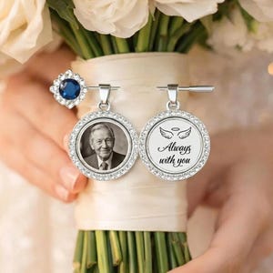 May include: A silver-toned bouquet charm set featuring a round photo pendant of an older man, a round pendant with the text Always with you and angel wings, and a small blue rhinestone accent, all attached to a white ribbon-wrapped bouquet handle.