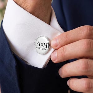 Op de afbeelding: Een ronde zilverkleurige metalen manchetknop bevestigd aan een wit overhemdmanchet. De voorzijde van de manchetknop is voorzien van de zwarte tekst A&H gevolgd door de datum 15-02-2026. Het accessoire heeft een glad, gepolijst oppervlak.