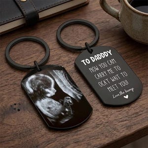 May include: Two black keychains with a tag shape. One keychain displays a sonogram image. The other keychain has the text "TO DADDY, NOW YOU CAM CARRY ME TO OCA'T WAIT TO MEET YOU! Love the bump" in white.