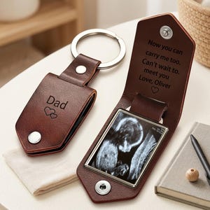May include: A brown leather keychain with the word "Dad" and two hearts engraved on it. The keychain opens to reveal a sonogram image. The text "Now you can carry me too. Can't wait to meet you Love, Oliver" is also engraved.