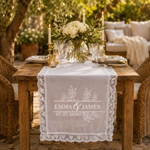 Puede incluir: Un camino de mesa de tela blanca con borde de encaje que presenta un diseño impreso de ramas con hojas, los nombres Emma & James y la fecha 07.27.2026. El camino está sobre una mesa de madera con un jarrón de cristal con rosas blancas y velas.