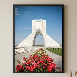 Puede incluir: Una impresión artística enmarcada que muestra la Torre Azadi en Teherán, un alto monumento de piedra blanca con un arco central, bajo un cielo azul claro. En primer plano, un parterre de jardín está lleno de rosas rojas y blancas en flor.