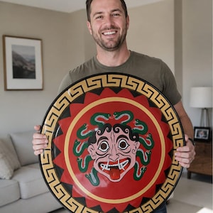 May include: A round shield featuring a Medusa design. The shield has a red center with a gold border and a black outer edge. The Medusa illustration has green snakes, a pink face, and a red tongue.