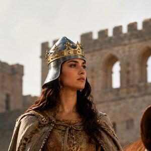 May include: A person wearing a metallic crown with a pointed design and a long, textured beige cloak over a gold-toned garment with intricate embroidery. The person is mounted on a brown horse, with a stone castle structure visible in the background.