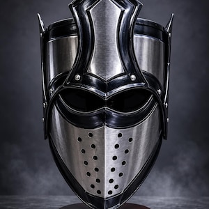 May include: A medieval-style helmet with a silver and black design. The helmet features a pointed top, a face guard with ventilation holes, and is mounted on a wooden base. The overall aesthetic is reminiscent of historical armor.