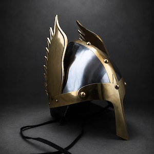 May include: A black and gold helmet with wing-like accents. The helmet features a polished black surface with gold-colored metal trim and decorative rivets. The wings are made of gold-colored metal and are attached to the sides of the helmet. Black ties hang from the bottom.