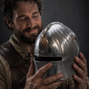 May include: A polished, silver-colored medieval helmet with a rounded top and a rectangular face opening. The helmet is held by a person with a beard and dark hair, against a dark background.