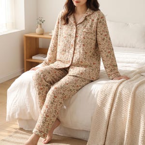 Op de afbeelding: Een tweedelige pyjamaset bestaande uit een shirt met lange mouwen en knoopsluiting en een bijpassende lange broek. De stof is lichtbeige met een herhalend bloemenpatroon in tinten rood, groen en bruin. Het shirt is voorzien van een kraag.