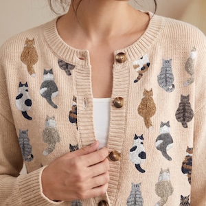 May include: A beige knit cardigan featuring a crew neckline and a front button closure with brown buttons. The garment is decorated with an all-over embroidered pattern of various cat breeds in different colours, including grey, brown, and black.