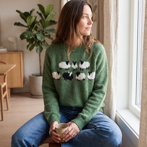 Puede incluir: Un suéter de punto de manga larga en color verde salvia con un patrón de intarsia repetitivo de pequeñas ovejas blancas y negras en el pecho. El suéter tiene un cuello redondo acanalado, puños acanalados y un dobladillo acanalado de tejido suave.