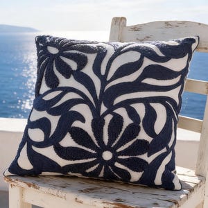 Könnte beinhalten: Ein quadratisches Zierkissen mit weißem Grund und einem dunkel marineblauen, gestickten Blumen- und Blattmuster. Das Kissen liegt auf einem weißen Holzstuhl mit abgenutzter Oberfläche und zeigt ein strukturiertes Design mit Reliefstickerei.