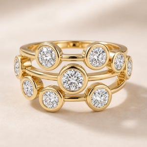 May include: A gold ring featuring a multi-layered band design with several round, bezel-set clear stones. The stones are arranged in a staggered pattern across the gold metal bands, creating a layered appearance with a polished, reflective finish.