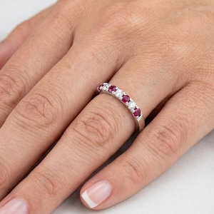 May include: A silver ring featuring alternating red and clear gemstones. The ring is worn on a finger, with the hand in the foreground. The gemstones are arranged in a line across the top of the ring.
