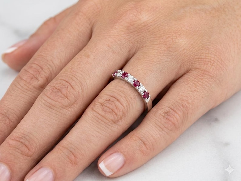 K&ouml;nnte beinhalten: Ein silberner Ring mit abwechselnd roten und klaren Edelsteinen. Der Ring befindet sich an einem Finger, wobei die Hand im Vordergrund ist. Die Edelsteine sind in einer Reihe &uuml;ber der Oberseite des Rings angeordnet.