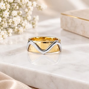 May include: A two-tone ring featuring a wide, polished gold-coloured band overlaid with a sleek, silver-coloured metal wave design that curves across the centre. The ring has a smooth, reflective finish and a distinct layered appearance.
