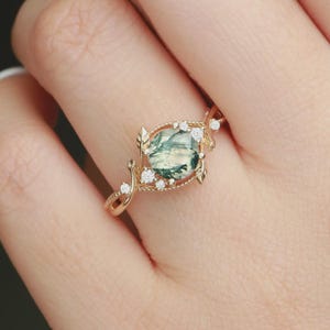 May include: A gold ring featuring a large, round, moss agate stone with green and white inclusions. The ring is adorned with small, clear gemstones and leaf-shaped accents. The band has a delicate, twisted design.