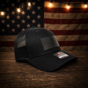 Op de afbeelding: Zwarte truckerpet met een mesh achterkant en een effen voorpaneel. De pet heeft een zwart leren embleem met een reliëf Amerikaanse vlag. De pet wordt getoond tegen een achtergrond van de Amerikaanse vlag en lichtslingers.