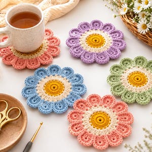 May include: Four round, flower-shaped crochet coasters in pastel pink, lavender, light blue, and sage green. Each coaster features a yellow center surrounded by a cream-colored ring and scalloped petals. A ceramic mug sits on the pink coaster.