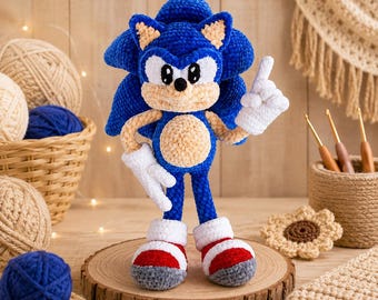 Könnte beinhalten: Ein handgefertigtes Häkel-Plüschtier der Figur Sonic the Hedgehog aus leuchtend blauem Garn mit beigem Bauch und Schnauze, weißen Handschuhen sowie rot-weißen Turnschuhen. Die Figur steht aufrecht mit einem nach oben zeigenden Finger.