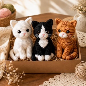 May include: Three crocheted cat figurines sitting in a cardboard box. One cat is solid white, one is black with a white chest and paws, and one is orange tabby with stripes. Each cat has dark plastic eyes, a pink stitched nose, and white whiskers.