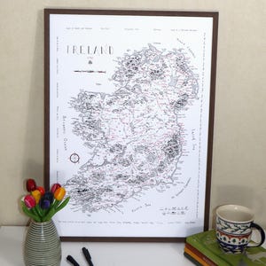 May include: A framed map of Ireland with detailed black and red markings. The map is in a dark brown frame and is displayed on a white surface. A vase of colorful tulips, pens, a mug, and books are also on the surface.