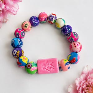 May include: A bracelet composed of round beads featuring various floral patterns in shades of pink, blue, purple, green, and yellow, centered with a rectangular pink bead embossed with Chinese characters, arranged in a circular formation.