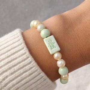 May include: A stretch bracelet featuring alternating round pale green and iridescent cream-colored beads. A rectangular light green mahjong tile bead with a green engraved character is centered among the beads, creating a cohesive design.
