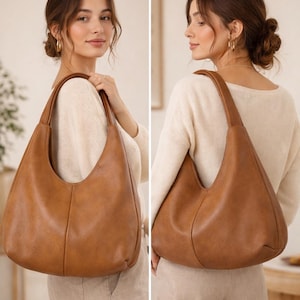 May include: A large, brown leather shoulder bag with a curved design. The bag has a single strap and is held by a person. The bag is a warm, medium brown colour and appears to be made of soft leather. The bag is a stylish accessory.