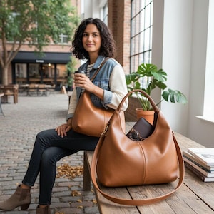 Puede incluir: Un bolso de hombro de cuero marrón con un asa superior y una correa larga ajustable. El bolso tiene una forma curva y un interior espacioso. Una segunda bolsa es visible en el fondo. Las bolsas están sobre una superficie de madera.