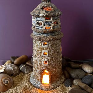 Pode incluir: Uma escultura de farol em cerâmica com um acabamento texturizado e desgastado em tons de castanho e cinzento. A estrutura apresenta várias pequenas aberturas quadradas e uma porta em arco, emitindo uma luz laranja quente do seu interior.