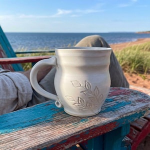 Peut inclure: Une tasse en céramique blanche avec un corps arrondi et une anse incurvée, présentant un motif de feuilles botaniques en relief sur la surface avant. La tasse a une finition mate lisse et un bord légèrement évasé, au design simple.