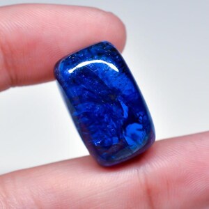 May include: A polished, rectangular gemstone with a deep blue color and swirling patterns. The stone is held between fingers, showcasing its glossy surface and natural variations. The gem's shape is slightly rounded, reflecting light.