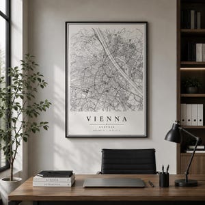 Könnte beinhalten: Eine gerahmte Schwarz-Weiß-Karte von Wien, Österreich, hängt an einer Wand über einem Schreibtisch. Die Karte ist detailliert und zeigt Straßen und Gebäude. Das Wort "WIEN" ist darunter gedruckt. Eine schwarze Schreibtischlampe und Bücher liegen auf dem Schreibtisch.
