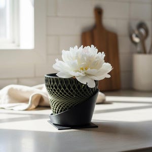 Könnte beinhalten: Eine weiße Pfingstrosenblüte steht in einem zweifarbigen Pflanzgefäß. Der obere Teil weist ein olivgrünes, komplexes Gitterdesign auf, während die untere Basis aus einem soliden, glatten schwarzen Material mit quadratischer Bodenplatte besteht.