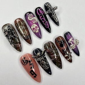 May include: A set of ten stiletto-shaped press-on nails featuring diverse designs including silver metal charms like skulls, crosses, and angels, alongside patterns of currency, marble textures, cherries, and black gothic lettering on purple, pink, and brown bases.