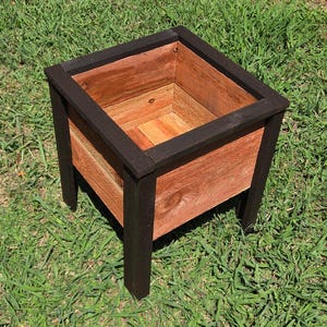 May include: A square wooden planter box featuring natural reddish-brown wood grain panels framed by a dark black painted border and four sturdy black legs. The container has an open top and a deep interior, showcasing a contrast between the dark frame and the warm wood tones of the side panels.