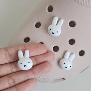 May include: White bunny shoe charms with black facial details, including a small cross for the mouth. The charms are shown on a light pink shoe and in a person's hand. These charms are a fun accessory for shoes.