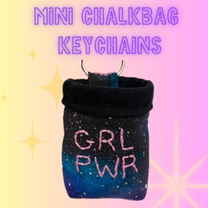 May include: A mini chalk bag keychain featuring a galaxy print in shades of blue, purple, and black. The fabric has the text GRL PWR embroidered in pink thread on the front. A black fabric rim surrounds the opening with a silver metal ring attached.