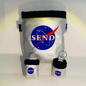May include: A silver chalk bag with a black rim and a blue circular logo featuring white stars, a red swoosh, and the word SEND in white capital letters. Two smaller matching silver and black pouches with key rings are positioned in front of the bag.