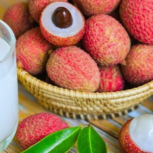 May include: A wicker basket overflowing with lychees, their red, textured skins contrasting with the white flesh of a partially peeled fruit. Green leaves and a glass of white liquid are also in view.