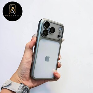 Może przedstawiać: Srebrna i przezroczysta obudowa iPhone'a z osłoną obiektywu aparatu. Obudowa ma logo Apple z tyłu i logo Aura Cases w lewym górnym rogu.
