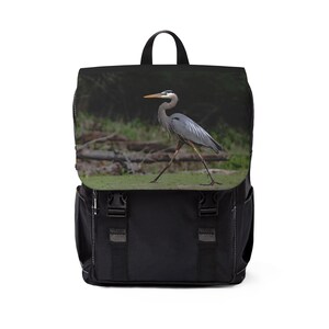 May include: A black backpack featuring a flap with a photographic print of a grey heron walking through a grassy area. The bag includes a top handle, two vertical straps with black plastic buckles on the front, and side pockets for storage.