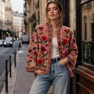 Könnte beinhalten: Eine altrosa Jacke mit lebendiger Blumenstickerei in Rot-, Rosa- und Grüntönen. Die Jacke hat einen geraden Schnitt, lange Ärmel und einen Stehkragen. Sie wird über einem weißen Tanktop und einer hellblauen Jeans aus Denim getragen.