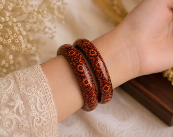May include: Two matching bangle bracelets featuring a glossy finish and a repeating intricate pattern in shades of deep red, burnt orange, and black. The bangles have a smooth, rounded profile and are worn together on the wrist.